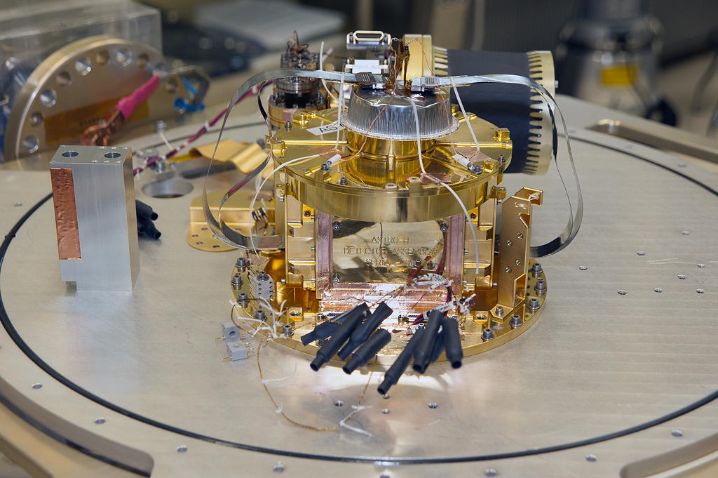 The gold-colored XRISM Resolve X-ray calorimeter assembly on a laboratory workbench, surrounded by wiring and test equipment.