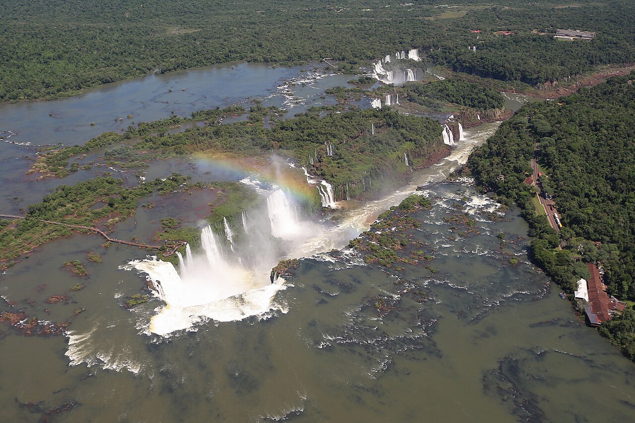 Aerial view of Iguazú Falls, with forested islands, river channels and a rainbow above the cascades.