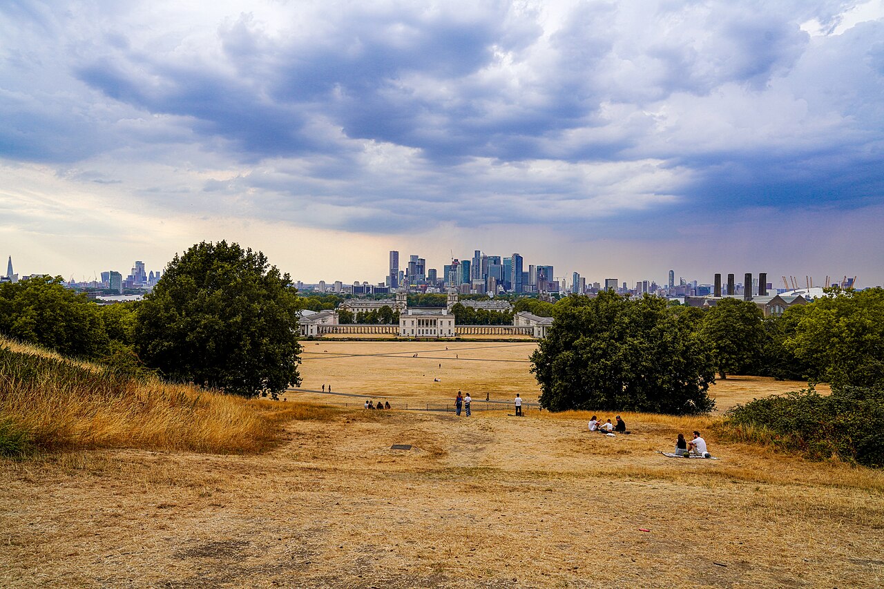 Scorched grass in Greenwich Park, London, with people, trees, and the city skyline beneath storm clouds.