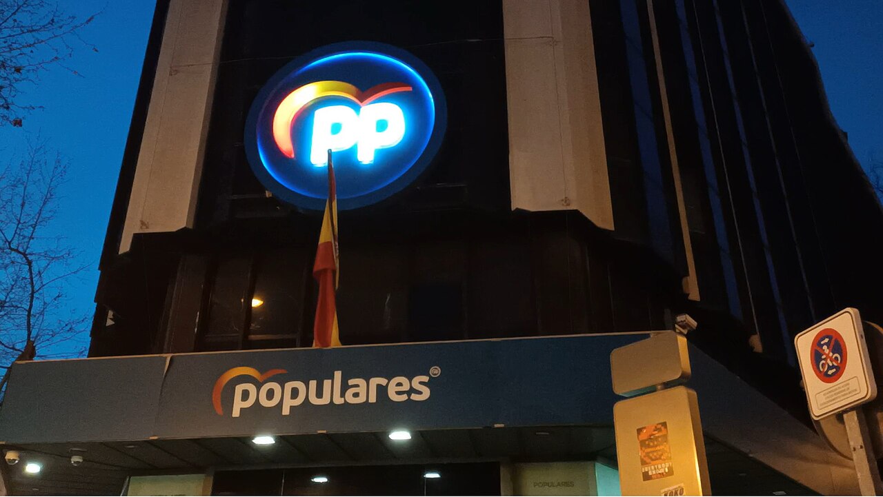 Illuminated PP logo above a Spanish flag and blue “populares” sign on the party’s Madrid headquarters at dusk.
