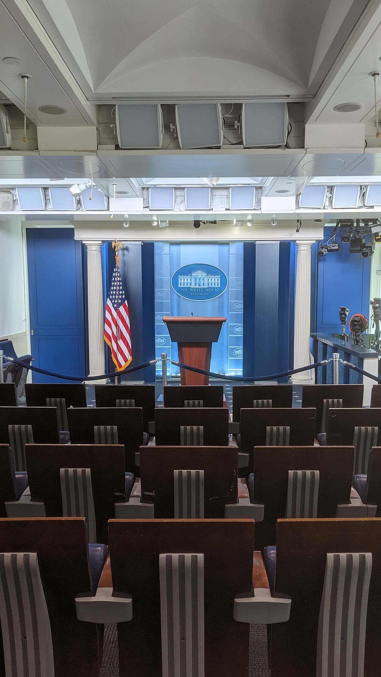 Empty James S. Brady Press Briefing Room with rows of seats facing the White House lectern and American flag.