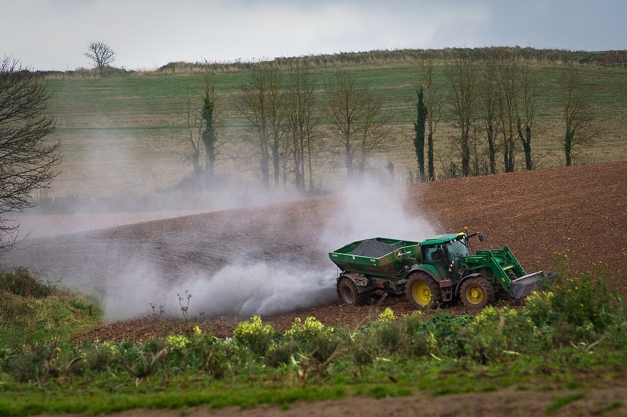 A tractor spreads a cloud of agricultural lime across a ploughed field bordered by trees.