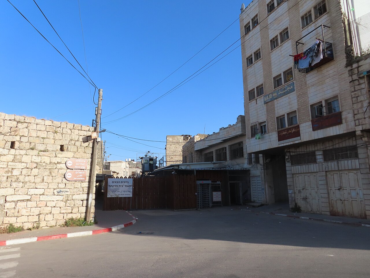 Abu Rish checkpoint in Hebron, with a metal barrier and pedestrian turnstile between stone buildings.