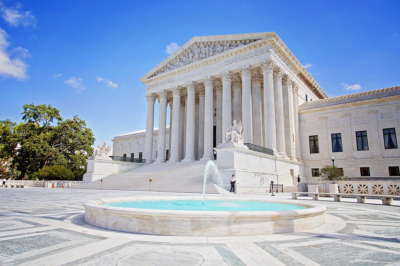 U.S. Supreme Court building with marble columns, broad steps and a fountain under a blue sky.