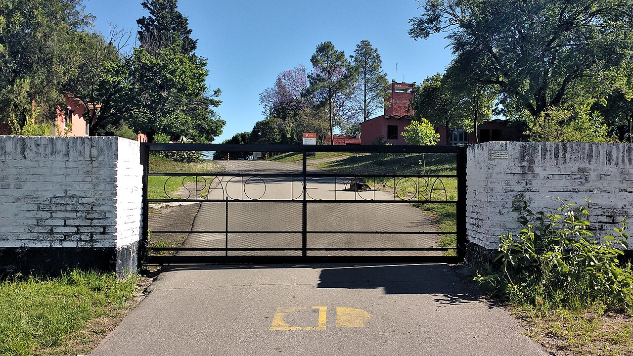 Black metal entrance gate between white brick walls at La Perla, with trees and buildings beyond.